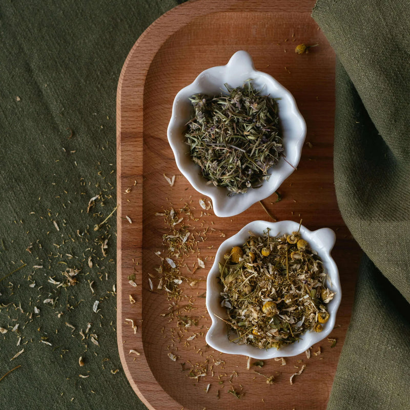 Two bowls of dried herbs on a wooden tray and green fabric.