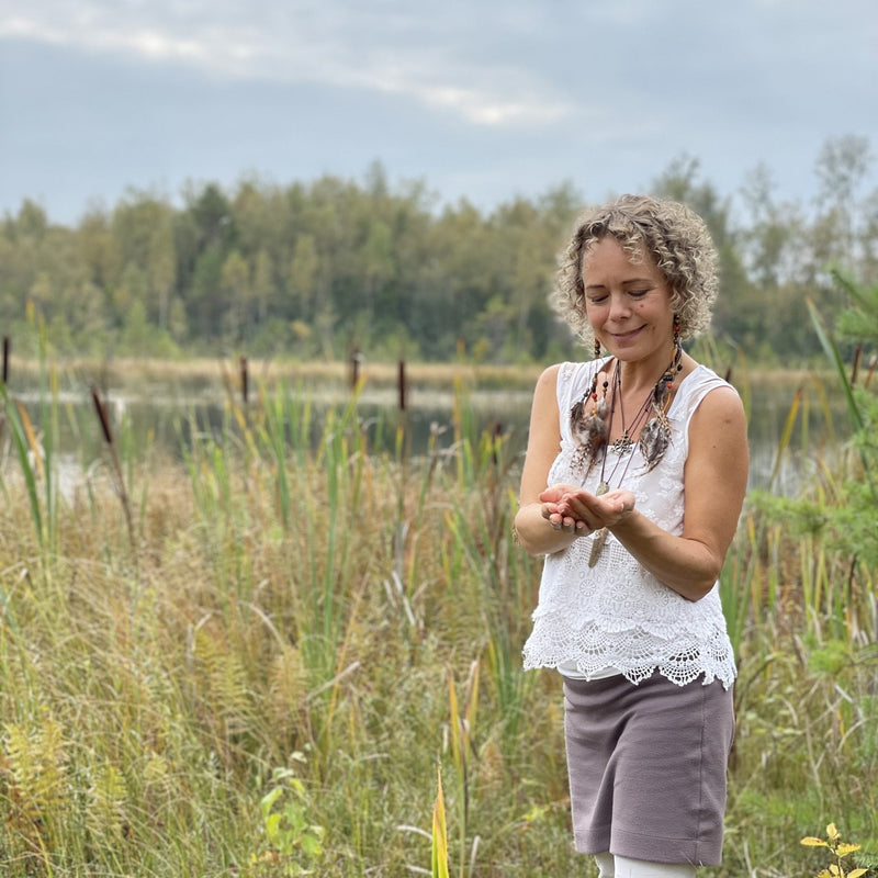A woman standing beside a pond in a meadow, looking down at her open hands.